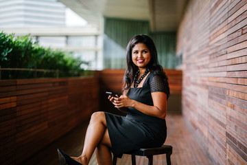 A confident and attractive Indian Asian woman is typing and using her smartphone as she sits on a chair outdoors during the day.
