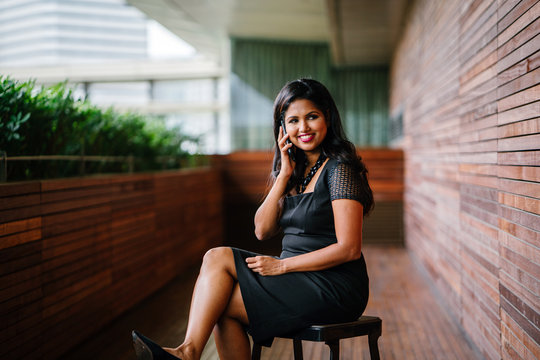 A Confident And Attractive Indian Asian Business Woman Is Speaking On Her Smartphone During The Day As She Sits On A Chair. She Is Smiling As She Talks On Her Phone.