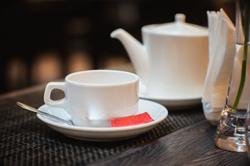 White empty cup and spoon for tea and red tea bag with teapot in a background