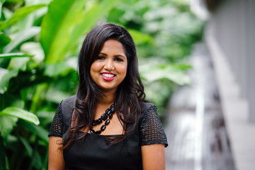 Portrait of a young Indian Asian business woman posing in front of lush green plants. She smiles happily in a black dress.