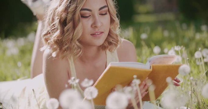 beautiful Asian girl surrounded by dandelions in the Park on a Sunny day with a book, having fun slow motion