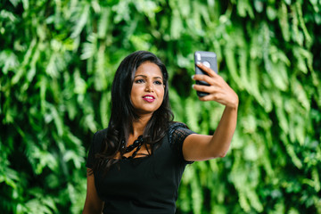 Portait of a young Indian Asian woman taking a selfie of herself using her mobile phone. She looks elegant and classy in her black necklace and dress.