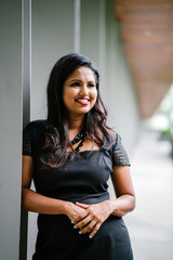 An Indian Asian professional woman leaning on a pillar while standing along the corridor on a sunny day. She is smiling confidently and looks dignified in her black dress.