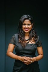 Portrait of a confident Indian woman in a studio.  She is wearing an elegant and professional black dress and is striking a classy pose.