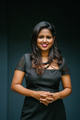 A portrait of a confident Indian woman in a studio. She is dressed in an elegant and professional black dress and is smiling for her head shot.