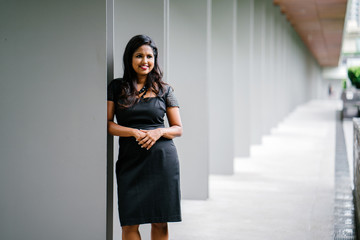 Portrait of a confident and mature Indian woman standing in a walkway and leaning against a pillar while striking an elegant pose. She looks very dainty in her black dress.