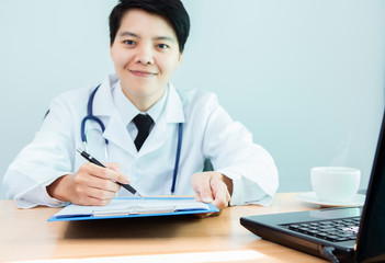 Asian young lesbian in Uniform of doctor Smiling and writing a patient report with digital technology laptop on the office desk.