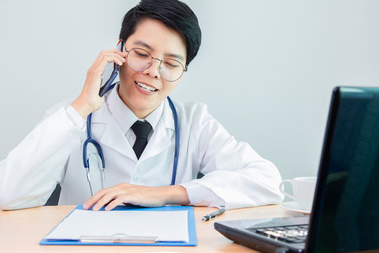 Asian Young Lesbian In Uniform Of Doctor Smiling Talking To A Counselor With Heart Disease. With Technology Mobile Phone Digital On The Office Desk.