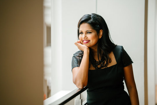 A successful Indian executive standing in the hallway and enjoying her view during the day. She is wearing an elegant dress and smiling while in front of the camera.