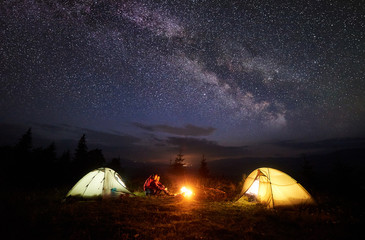 Fototapeta na wymiar Night camping in mountains. Bright campfire burning between two hikers, man and woman sitting opposite each other in front of illuminated tents under beautiful evening starry sky and Milky way