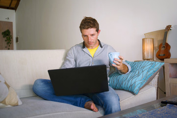 young attractive and successful man working from home living room sitting on couch networking on laptop computer drinking coffee happy in freelancer concept