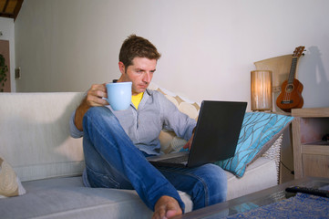 young attractive and successful man working from home living room sitting on couch networking on laptop computer drinking coffee happy in freelancer concept