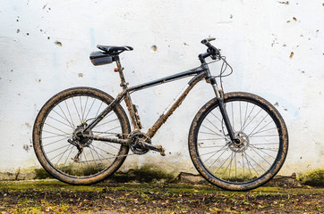 Fototapeta na wymiar Dirty Mountain Bike Covered with Mud After Riding in Bad Weather Stands. Grey 29er Hardtail Bike on White Wall Background