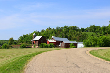 Fototapeta premium Agriculture and farming background. Rural life concept.Scenic countryside spring landscape with rural road along farm buildings and ted barns on a woods background. Midwest USA,Wisconsin,Madison area.