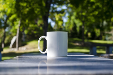 A long view of a blank white coffee mug on the marble bench of the park. 
