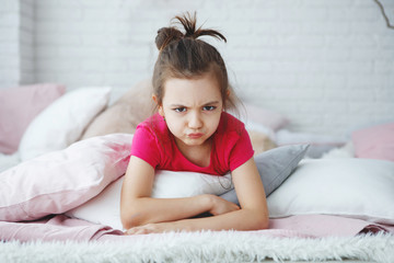 A young long-haired girl in a pink T-shirt lies in bed with cushions scowling and looks suspiciously at the camera