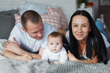 Obraz premium Young mom and dad and their baby son lie in bed and look at the camera indoors. The concept of a happy family
