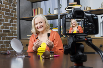 Progressive modern granny concept. Elderly woman blogger wearing colorful clothes recording her doing make up online video with camera, holding brushes. Granny’s workplace with powder, brush, mirror