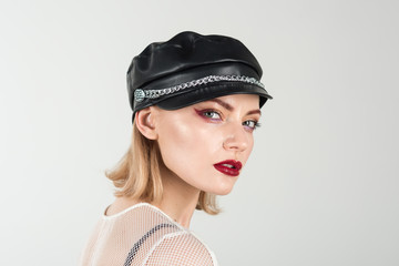 Trendy girl in rock style. Cool look of sexy girl with fashionable makeup. Rock woman with blonde hair on grey background. Girl in leather cap. Fashion model with stylish hairdo.