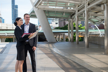 Asian business woman wear suit holding document and point her finger to building and discussing business future with Caucasian businessman at the place outside building in the city