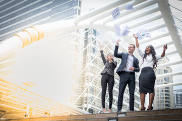 Caucasian businessman, Asian business woman and Black business woman throwing paper in the air and raising up hands to cheerful and celebrated for successful in career and mission.