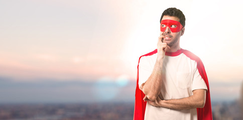 Superhero man with mask and red cape having doubts and with confuse face expression while looking up. Questioning an idea on a sunset background © luismolinero