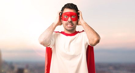 Naklejka premium Superhero man with mask and red cape takes hands on head because has migraine on a sunset background