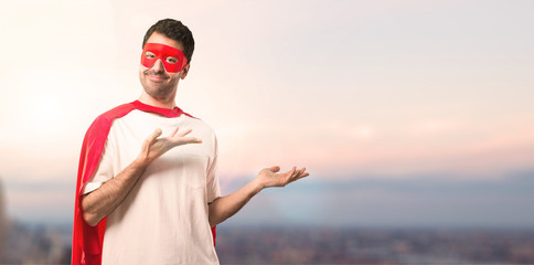 Superhero man with mask and red cape extending hands to the side and smiling for presenting and inviting to come on a sunset background