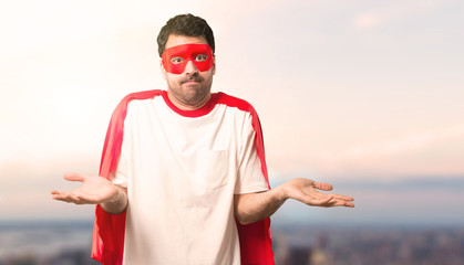 Naklejka premium Superhero man with mask and red cape making unimportant and doubts gesture while lifting the shoulders and the palms of the hands on a sunset background