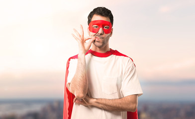 Superhero man with mask and red cape showing a sign of closing mouth and silence gesture doing like...