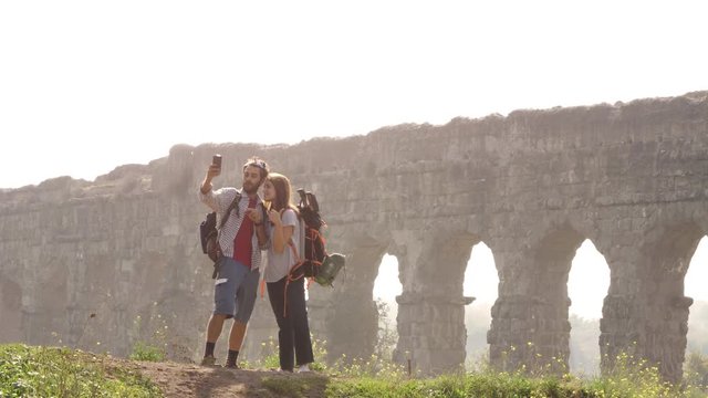 Young lovely couple backpackers tourists taking selfies photo on hill in front of roman aqueduct arches in parco degli acquedotti park ruins in rome on romantic misty sunrise with sleeping bag slow