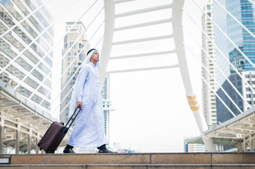 Muslim man wear white turban and dress walking with luggage in the city and modern building background. concept of business travel.