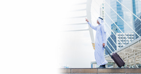 Muslim man wear white turban and dress walking with luggage in the city and pointing finger to building. concept of business travel and copy space for text.