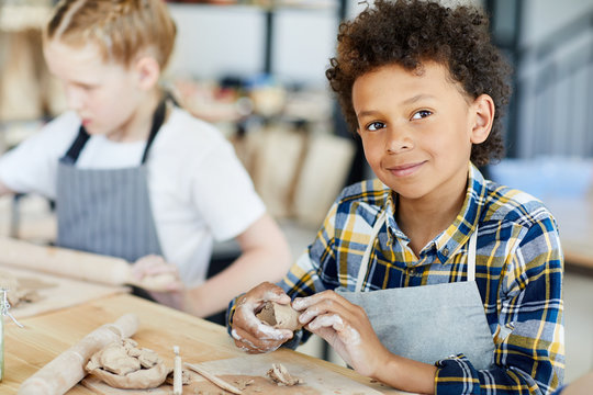 Creative schoolboy with piece of clay sitting by table in workshop and learning to make earthenware