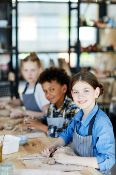 Youthful girl looking at camera with smile while working with clay at lesson on background of classmates