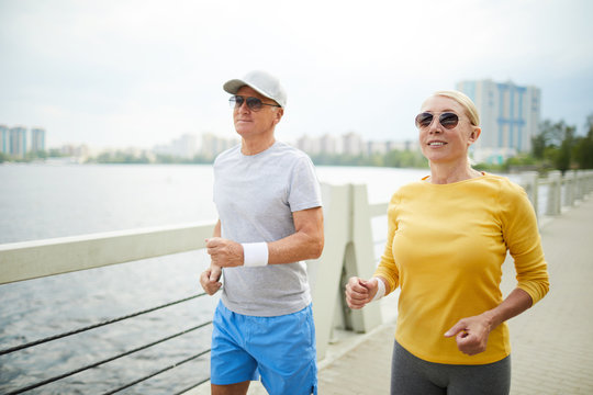 Two Active Seniors In Sportswear And Sunglasses Jogging By Waterside On Summer Morning