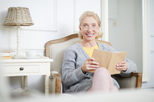 Smiling aged blond woman with book looking at camera while having rest at home