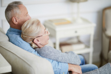 Mature man embracing his wife while relaxing on sofa in front of tv set in the evening