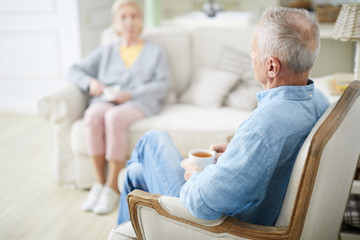 Senior man with cup of tea and his wife relaxing in living-room and having talk in the evening