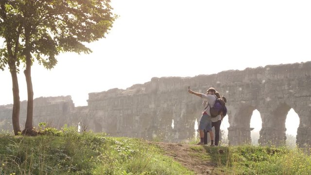 Young lovely couple backpackers tourists taking selfies photo on hill in front of roman aqueduct arches in parco degli acquedotti park ruins in rome on romantic misty sunrise with sleeping bag slow