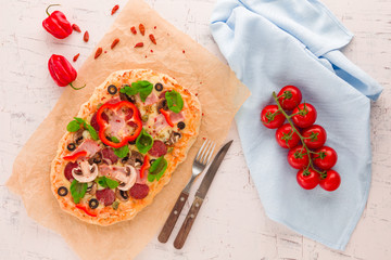 Homemade pizza with ham, pepperoni, mushrooms, capers and olives on a white concrete surface.