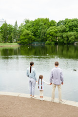 Little girl holding her parents by hands while family standing by waterside in modern park