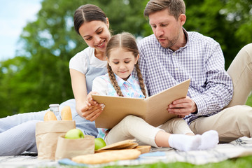 Two young parents and little daughter reading book while relaxing on ground in park