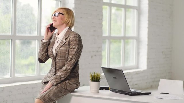 Confident businesswoman dressed in checked suit and eyeglasses talking via smartphone in wide white ofice room sitting on working table with laptop
