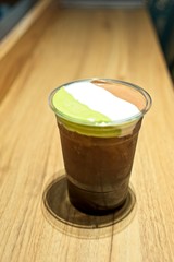 A plastic glass of iced tea with layer of cream cheese foam, Trendy food and drinks, Cheese Tea with three whipped cream cheese consists of matcha, chocolate and vanilla. on wooden table 