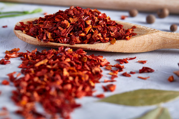 Red hot chillies pepper flakes in wooden spoon on white textured background, top view, close-up, selective focus.