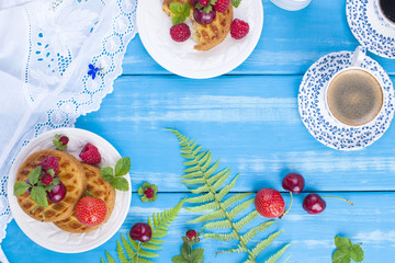 Small cupcakes with berries, strawberries and cherries. Delicious sweet breakfast and fragrant coffee, on a blue wooden background and flowers. Copy space