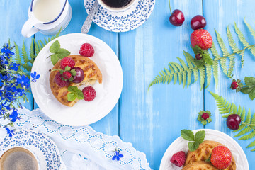 Small cupcakes with berries, strawberries and cherries. Delicious sweet breakfast and fragrant coffee, on a blue wooden background and flowers. Copy space. Above. flat lay