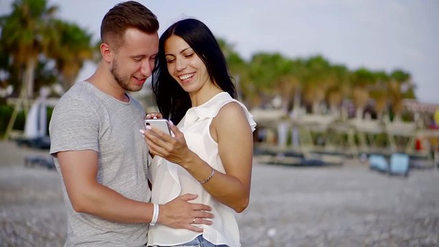 happy young couple looking at mobile phone screen standing on the beach