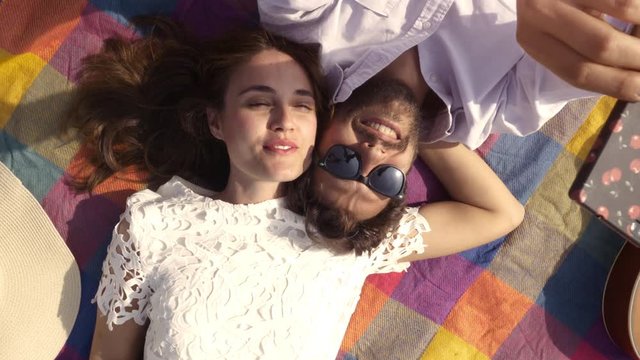 Happy lovely young couple lying on colorful blanket in the park taking selfies smartphone romantic with guitar sunglasses beautiful attractive girl top view rotating camera slow motion